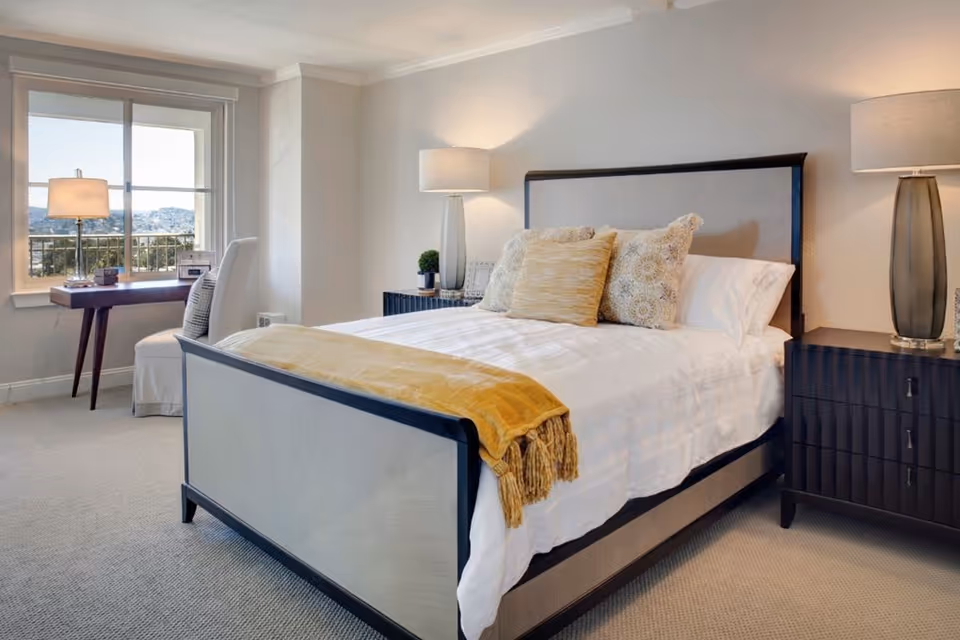 A neatly made bed with a light-colored headboard and footboard, adorned with decorative pillows and a yellow throw blanket. On either side of the bed are dark wooden nightstands with modern lamps. A window with a view of the outdoors is visible, along with a small desk and chair positioned near the window.