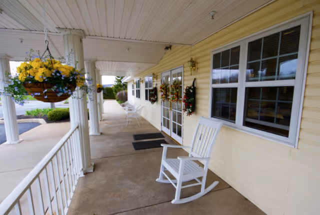 Covered porch area with white rocking chairs and hanging flower baskets, attached to a building with yellow siding and decorated with wreaths on the doors and windows.