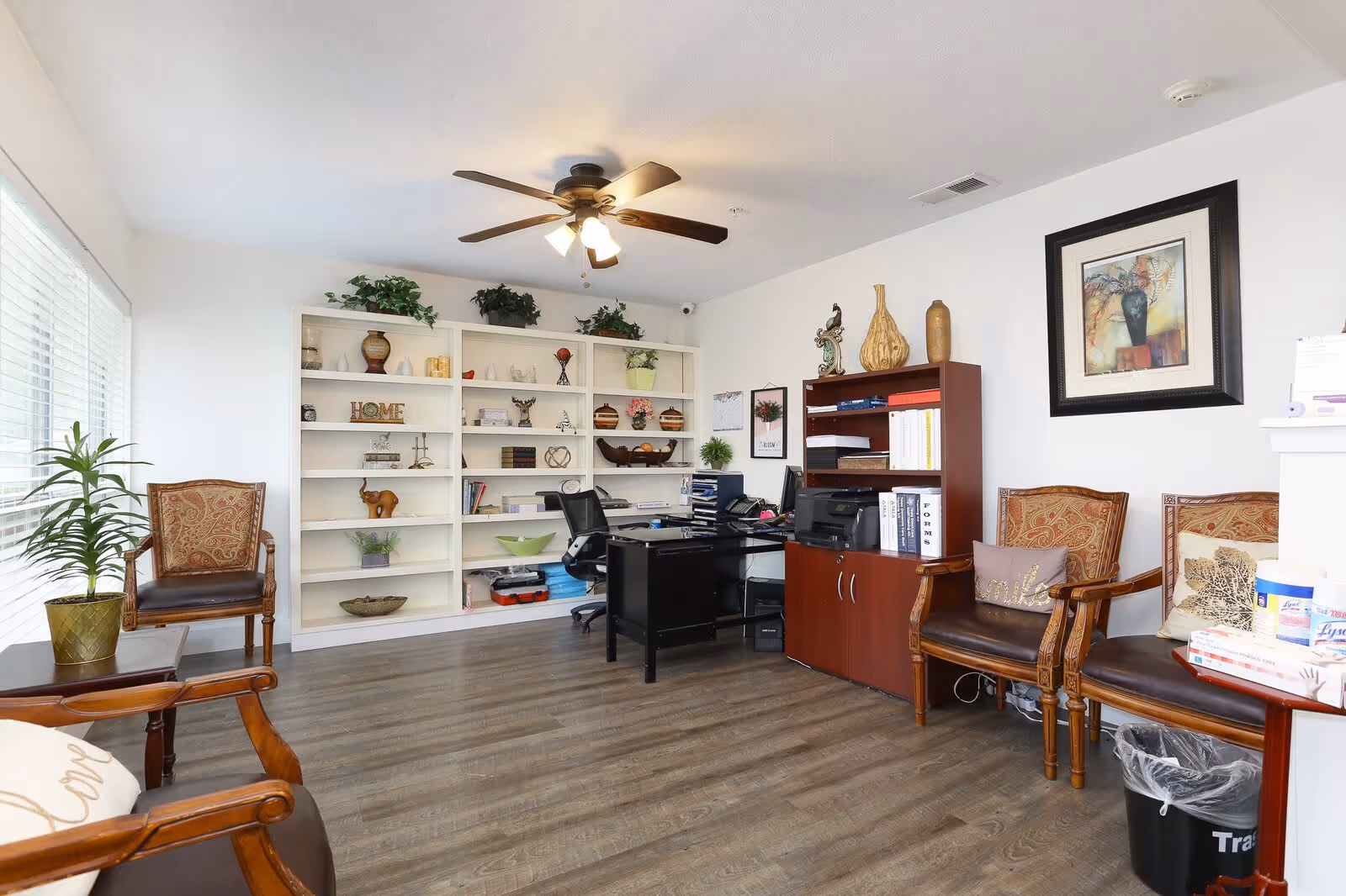 A bright office space with wooden flooring, a ceiling fan with lights, and white walls. The room features a black desk with office supplies and a computer, a wooden bookshelf filled with books and decorative items, and a large white shelving unit with various decorative pieces and plants. There are several wooden chairs with patterned cushions around the room, a small table with a potted plant, and framed artwork on the walls. A trash bin is visible near the chairs.