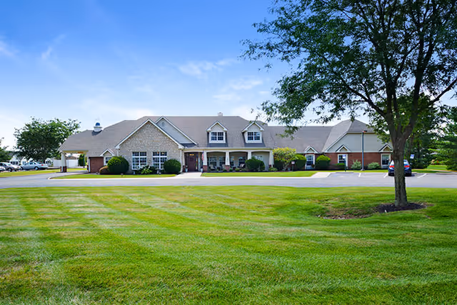 Front view of a single-story brick and siding senior living building with a wide manicured lawn, a tree, and a blue sky.