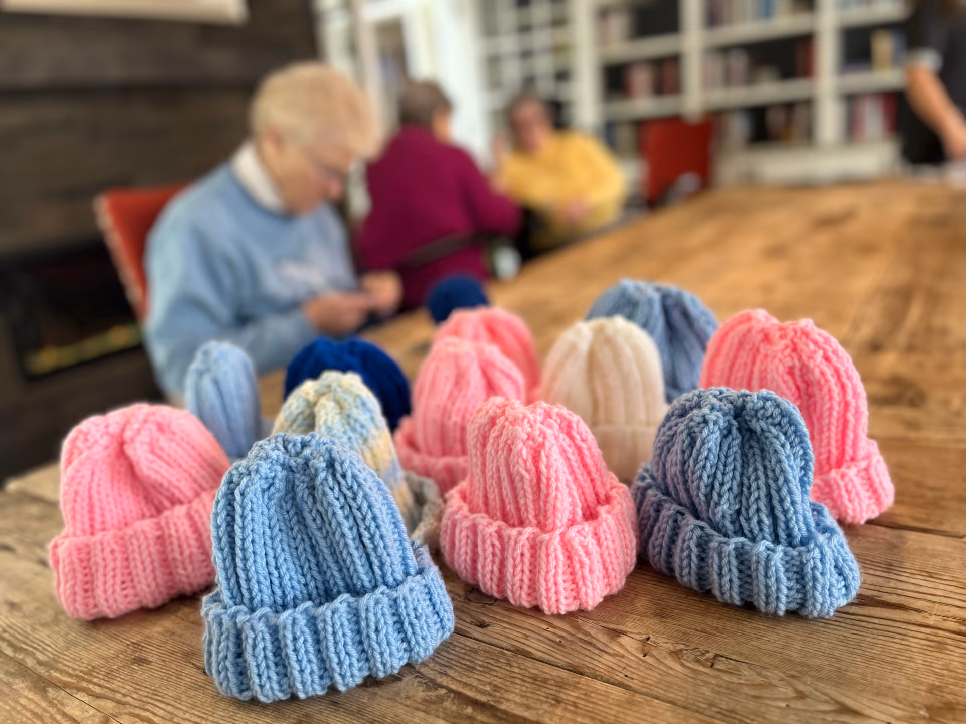 Several hand-knitted pastel baby hats arranged on a wooden table in a communal room with blurred residents and bookshelves in the background.