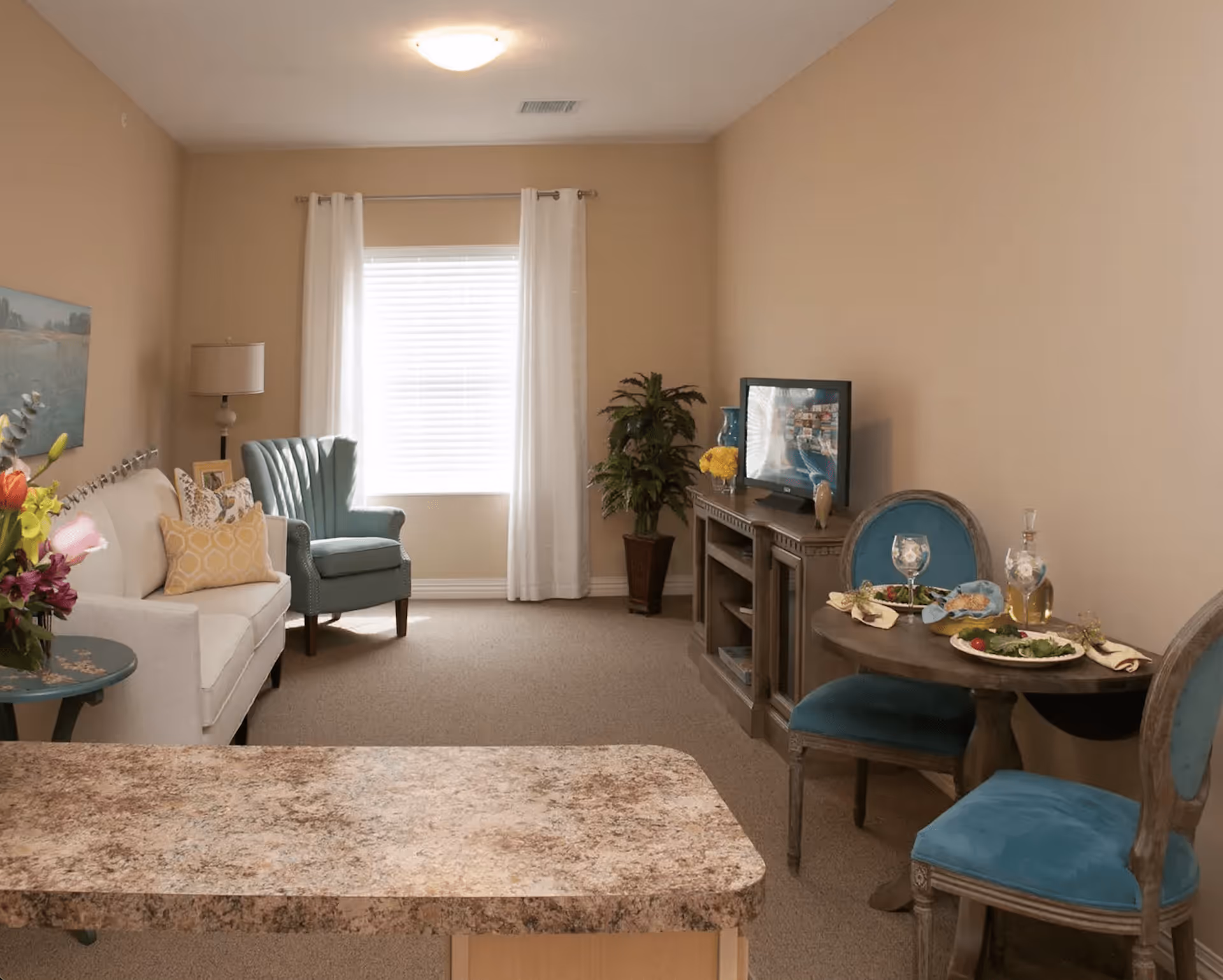A cozy living room area with a white sofa adorned with yellow and patterned pillows, a teal armchair, a small round table with two blue upholstered chairs set with plates of food and glasses, a TV on a wooden stand, a potted plant, and a window with white curtains letting in natural light.