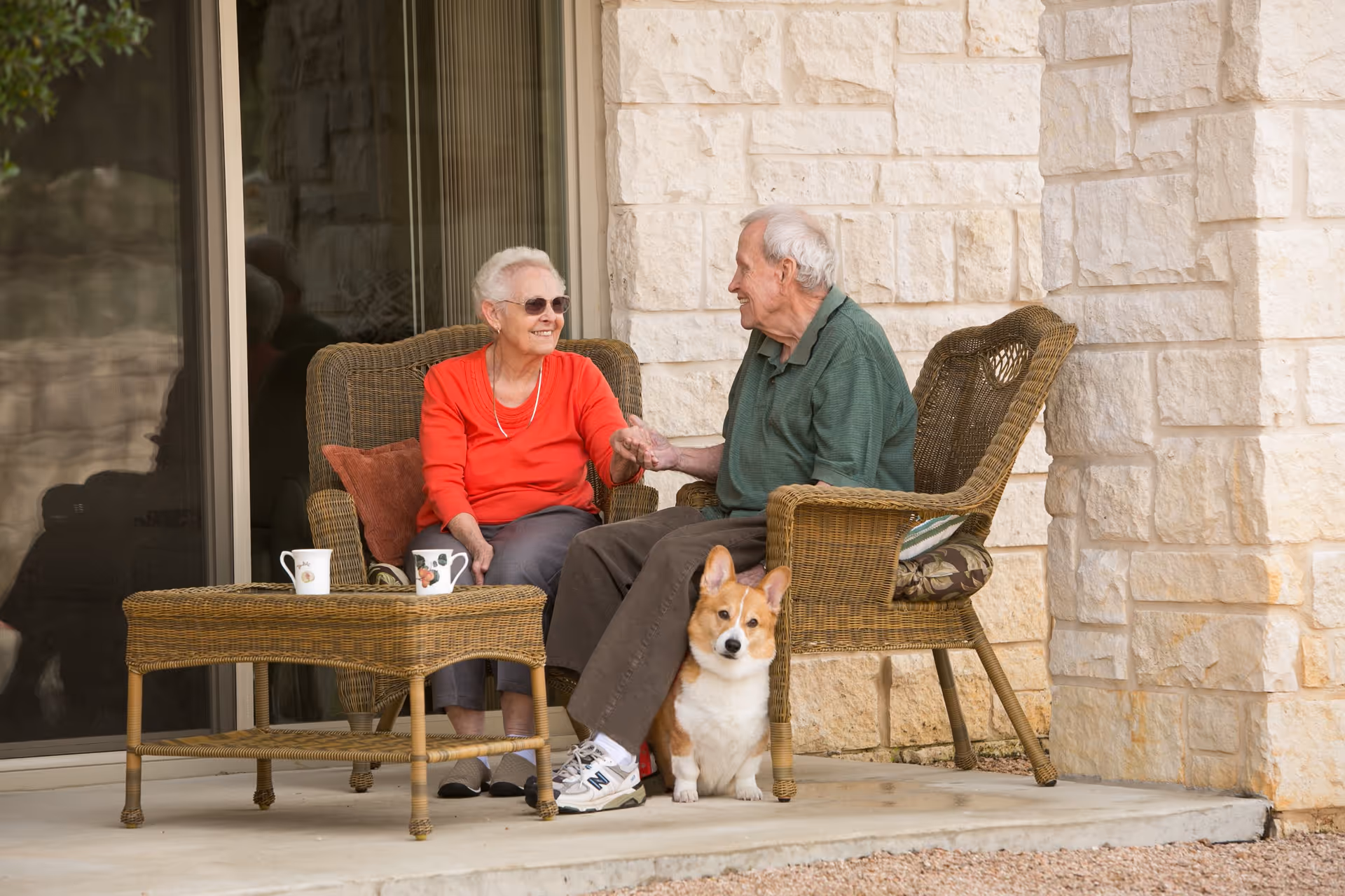 An elderly couple sitting on wicker chairs on a stone patio outside a building, holding hands and smiling at each other. A small dog sits on the ground between them. There is a wicker table with two mugs on it in front of them.