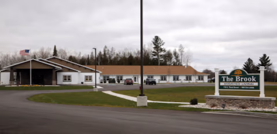 Front view of a single-story senior living building with a circular driveway, lawn, flagpole, and a sign reading 'The Brook'.