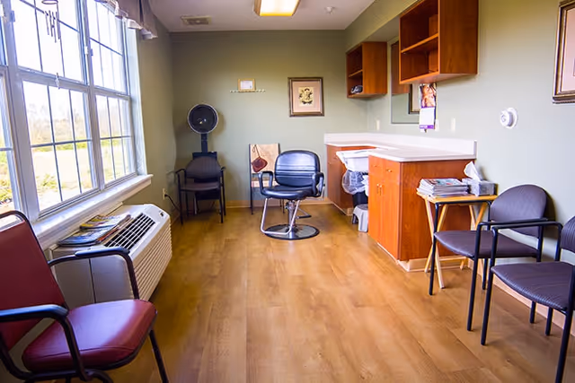 A small salon room with wooden flooring and light green walls. There is a large window on the left side letting in natural light. The room contains a black salon chair in the center, a hair dryer chair with a hood dryer, several waiting chairs along the walls, a countertop with a sink, and shelves mounted on the wall. Magazines are stacked on a small table next to the chairs.