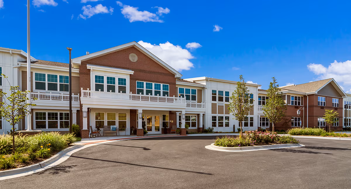 Front exterior view of a large, two-story senior living facility building with a combination of brick and white siding, multiple windows, a covered entrance with seating, landscaped greenery, and a clear blue sky with some clouds.