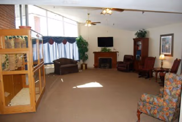 A spacious living room with beige carpet, a wooden fireplace with a TV mounted above it, several chairs including a floral armchair and a dark brown loveseat, a wooden display cabinet on the left, ceiling fans with lights, and large windows with blue curtains allowing natural light to enter.