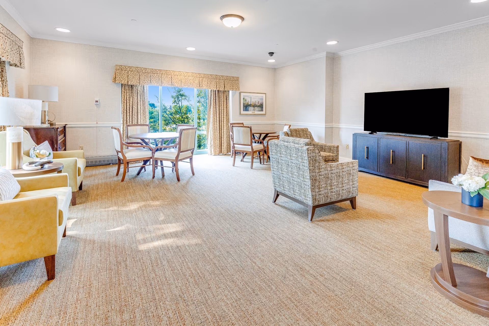 Bright communal living room with armchairs, a TV on a cabinet, and a round dining table by sliding glass doors.