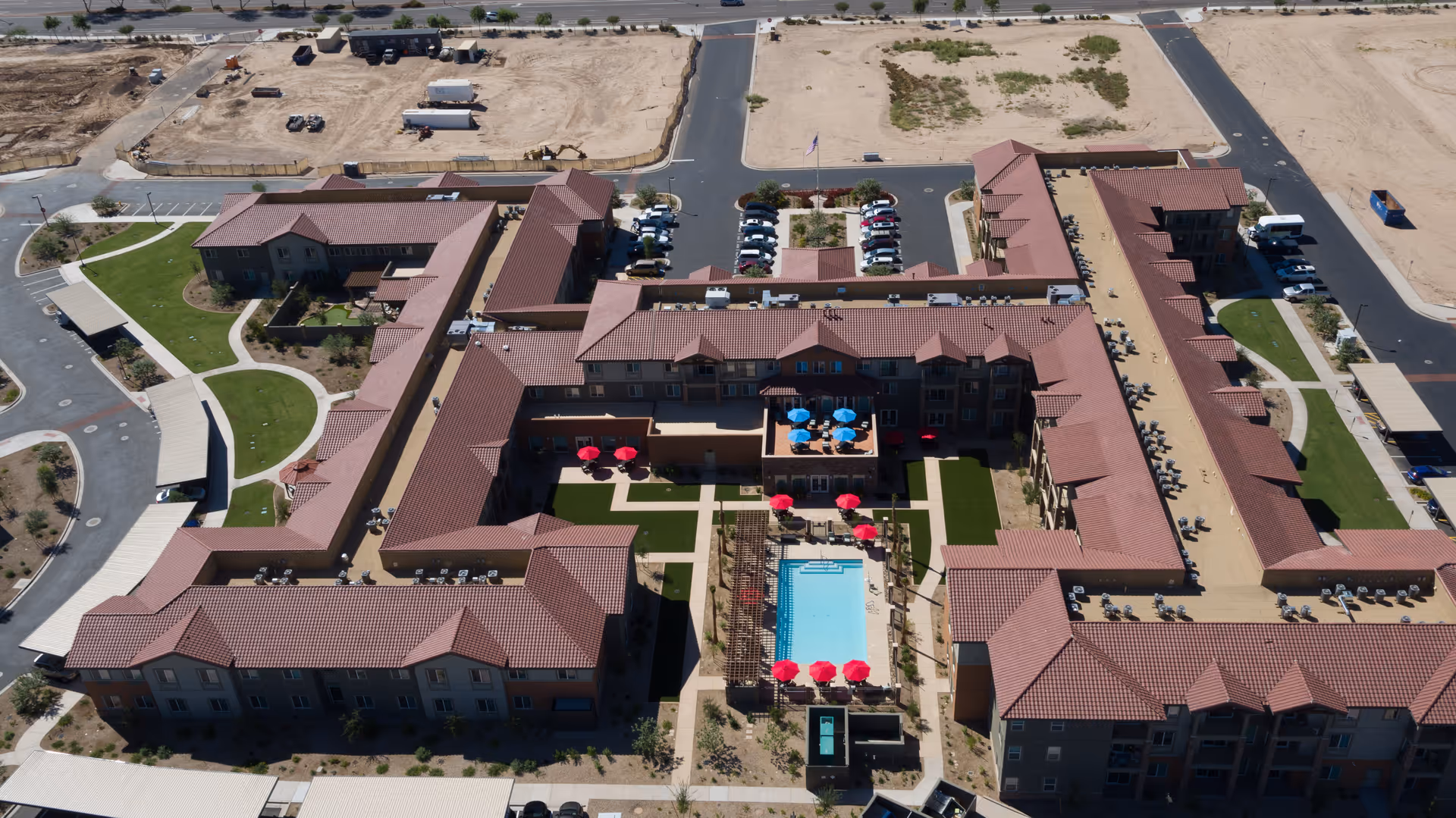 Aerial view of The Enclave at Gilbert Senior Living facility showing a large U-shaped building with red-tiled roofs surrounding a central courtyard. The courtyard features a swimming pool with red umbrellas, seating areas with blue umbrellas, and landscaped green spaces. Parking lots and roads surround the building, with some undeveloped land visible in the background.