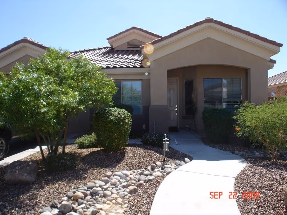 Front exterior view of a single-story residential building with a tiled roof, beige stucco walls, a small covered porch, and a pathway leading to the front door. The landscaping includes bushes, small trees, and rocks.