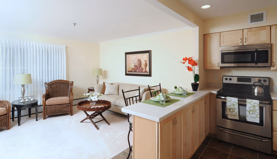 Open-plan living area and kitchen with a sofa, wicker chairs, coffee table, breakfast bar set for two, and stainless steel stove and microwave.