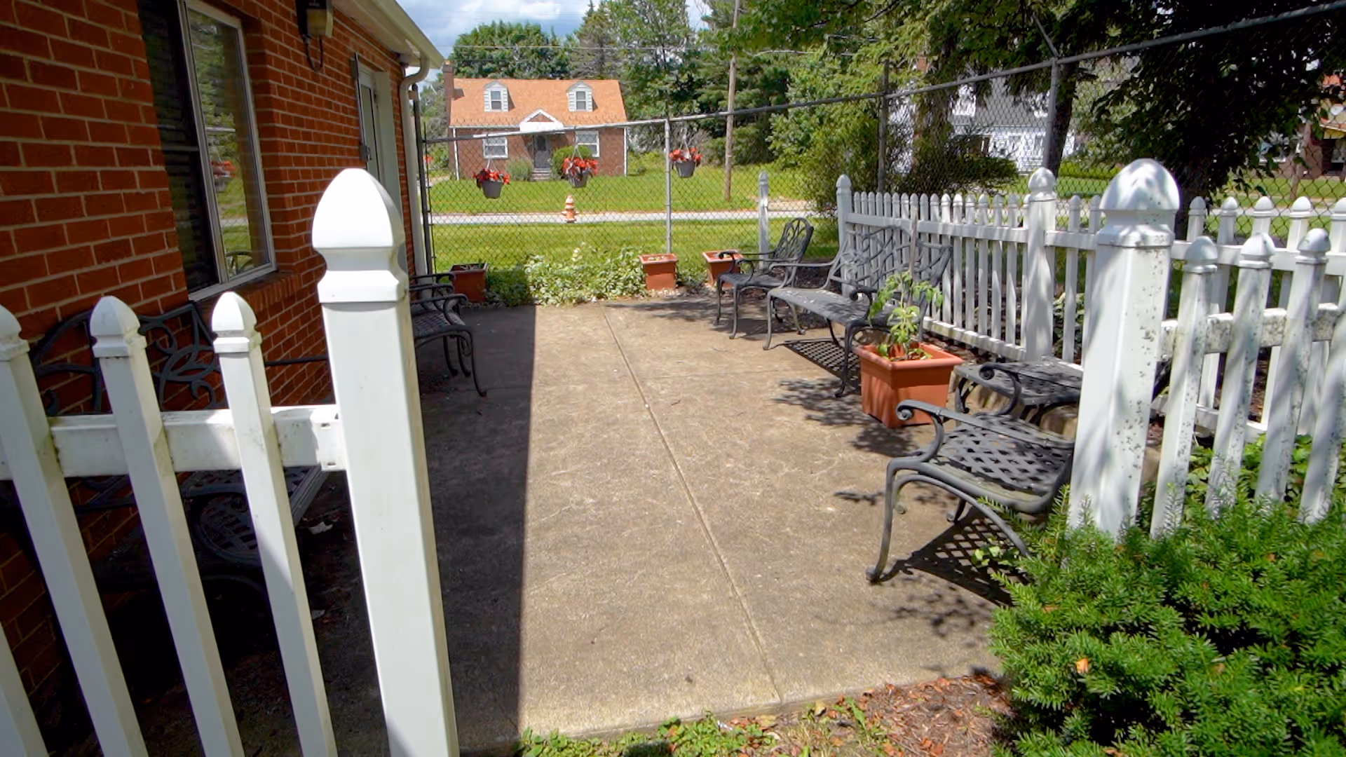 Outdoor patio area with a concrete floor, surrounded by white picket fencing and a brick building wall. Several black metal benches and chairs are placed along the sides, with potted plants and hanging flower baskets visible. Green bushes and a grassy area are also present.