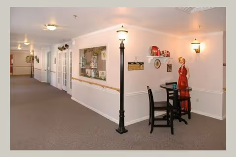 Interior hallway of a senior living facility with beige walls and carpeted floor. There is a small seating area with a black table and two chairs, a decorative lamp post, and a mannequin dressed in a red outfit. The hallway has wall-mounted lights and a window looking into another room.