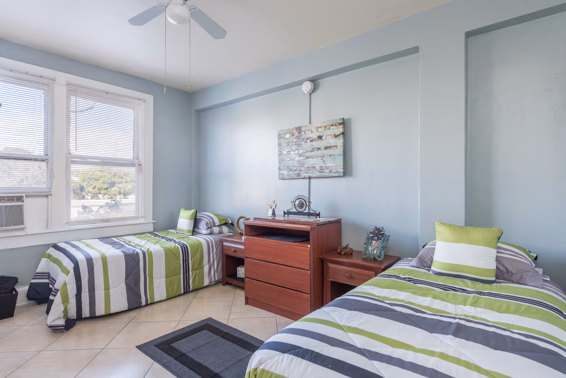 A bright bedroom with two single beds featuring striped green, gray, and white bedding. Between the beds is a wooden dresser with a clock and decorative items on top. The room has light blue walls, a ceiling fan, a window with blinds, and tiled flooring with a small rug.