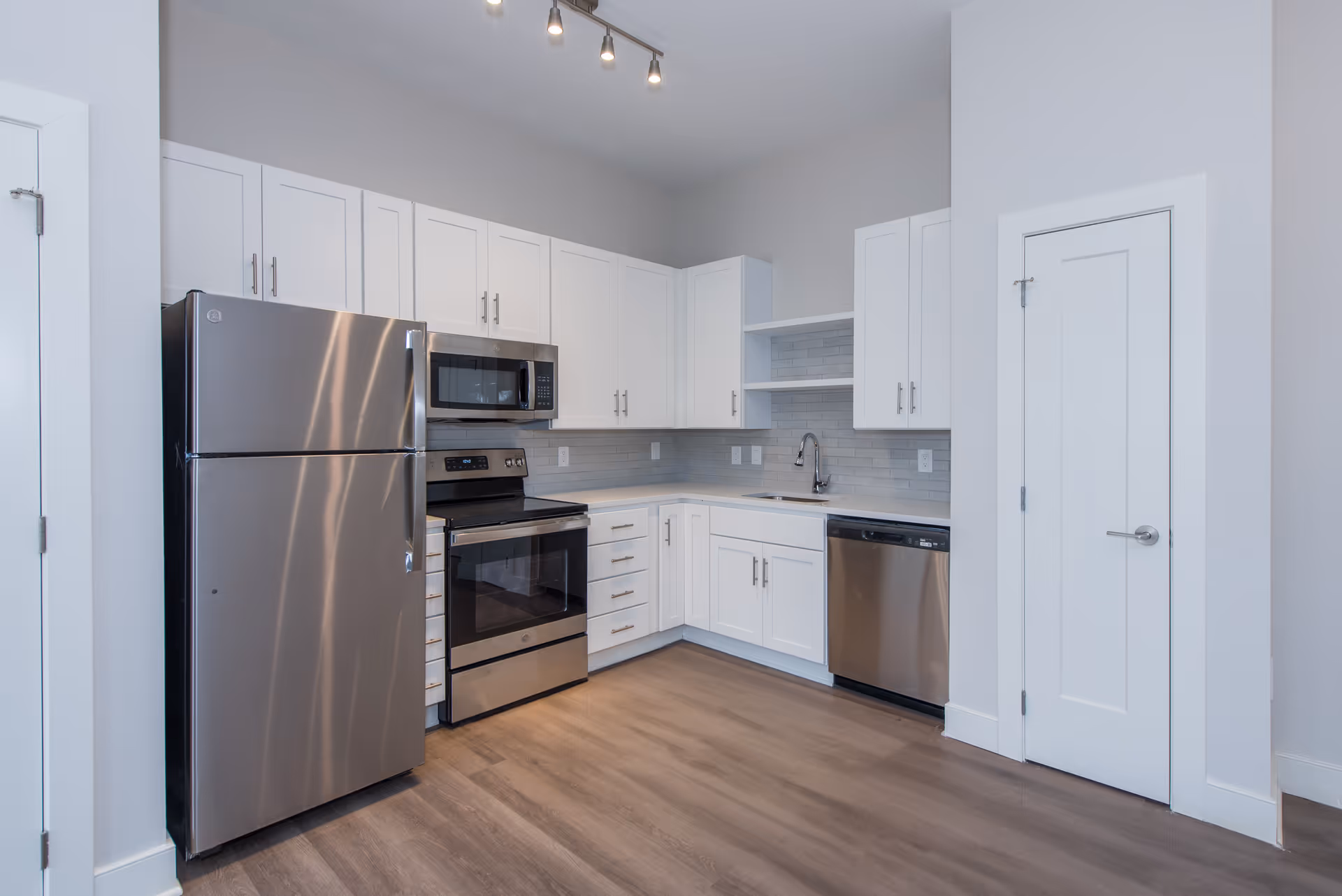 Modern kitchen with stainless steel refrigerator, oven, microwave, and dishwasher. White cabinets with silver handles, light gray tiled backsplash, and wood flooring.