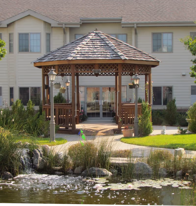 A wooden gazebo with a shingled roof stands in front of a beige building with multiple windows. The gazebo is surrounded by green shrubs, grass, and a small pond with water lilies and a small waterfall in the foreground. There are two lamp posts near the gazebo and a paved walkway leading to it.