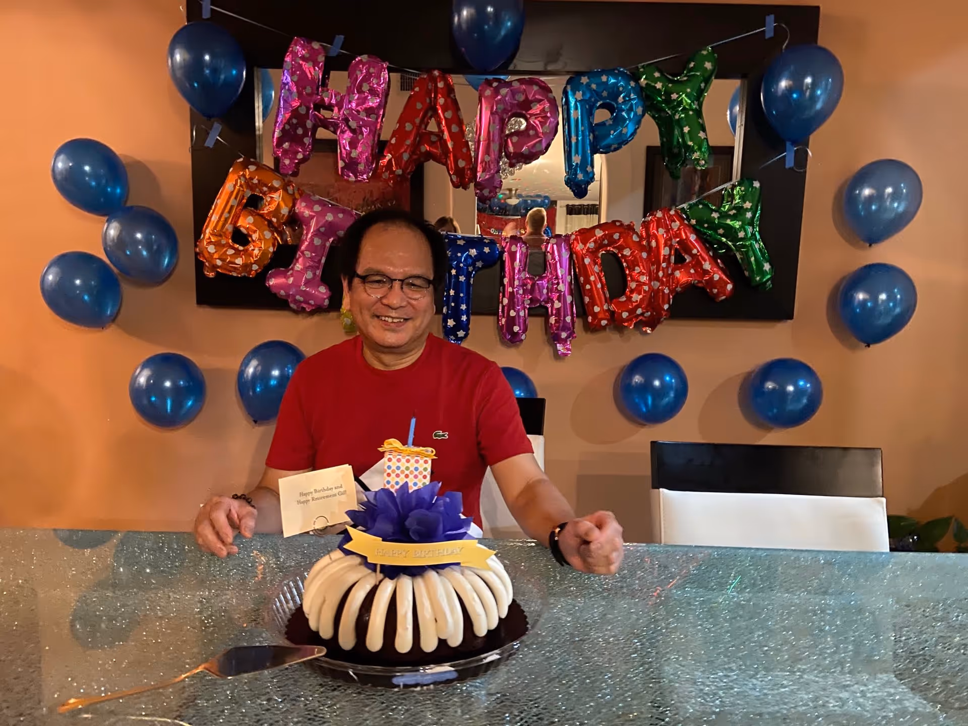 A man wearing glasses and a red shirt is sitting at a table with a birthday cake in front of him. Behind him, colorful balloons spell out 'HAPPY BIRTHDAY' and there are additional blue balloons decorating the wall.