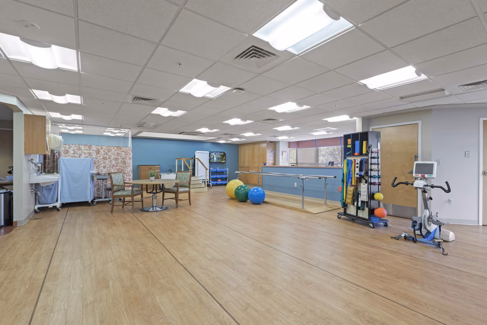 A spacious rehabilitation and therapy room with wooden flooring, exercise equipment including therapy balls, parallel bars, a stationary bike, and resistance bands. There is a small round table with chairs, a sink area, and a partition with floral curtains. The room has bright overhead lighting and large windows with blinds.