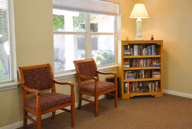 A cozy interior corner with two wooden armchairs upholstered in patterned fabric placed next to a window with blinds. Beside the chairs is a wooden bookshelf filled with books, topped with a lit table lamp and small decorative items. The room has beige walls and carpeted flooring.