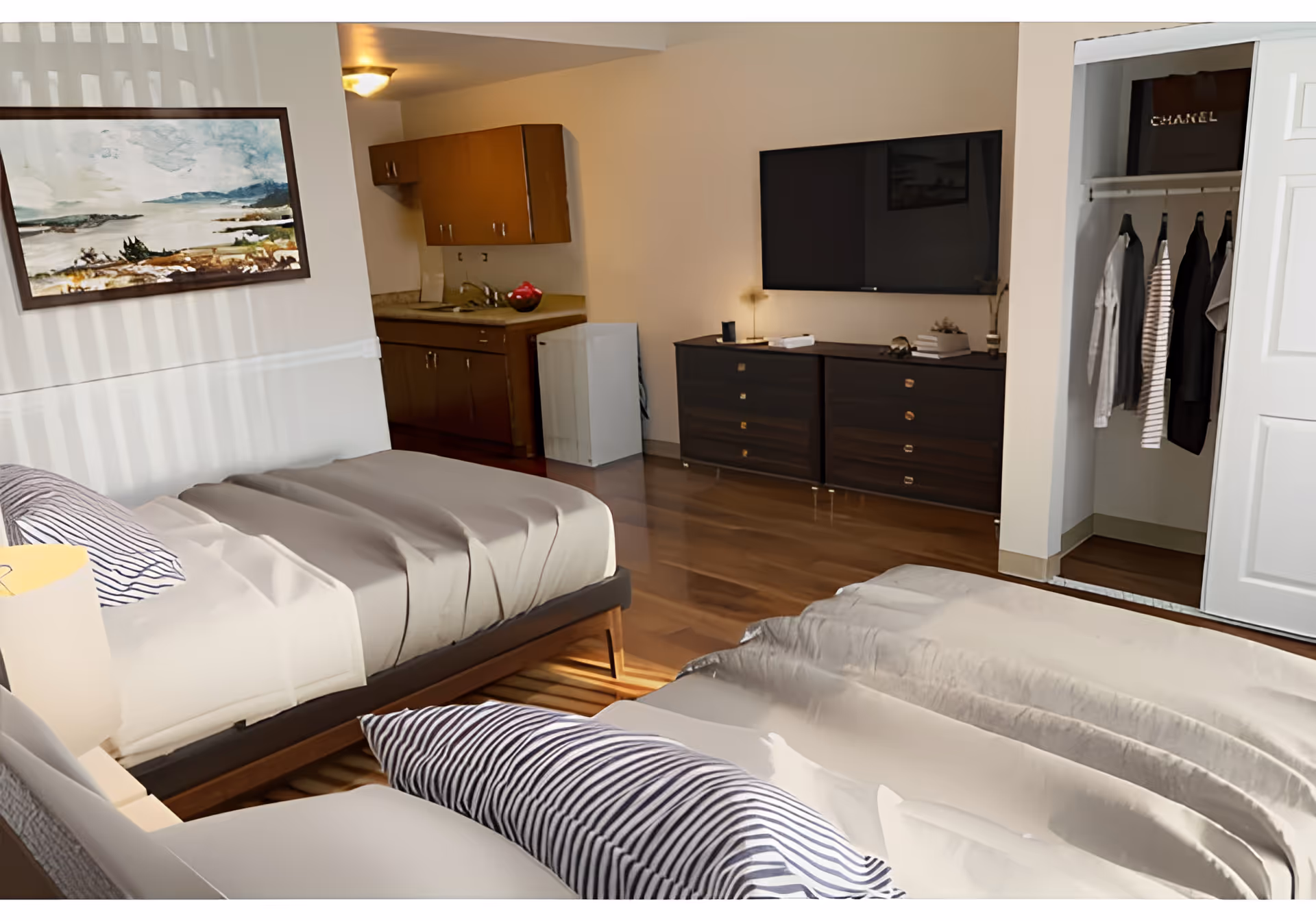 Studio-style bedroom with two beds, a wall-mounted TV over a dresser, a small kitchenette in the back and an open closet.