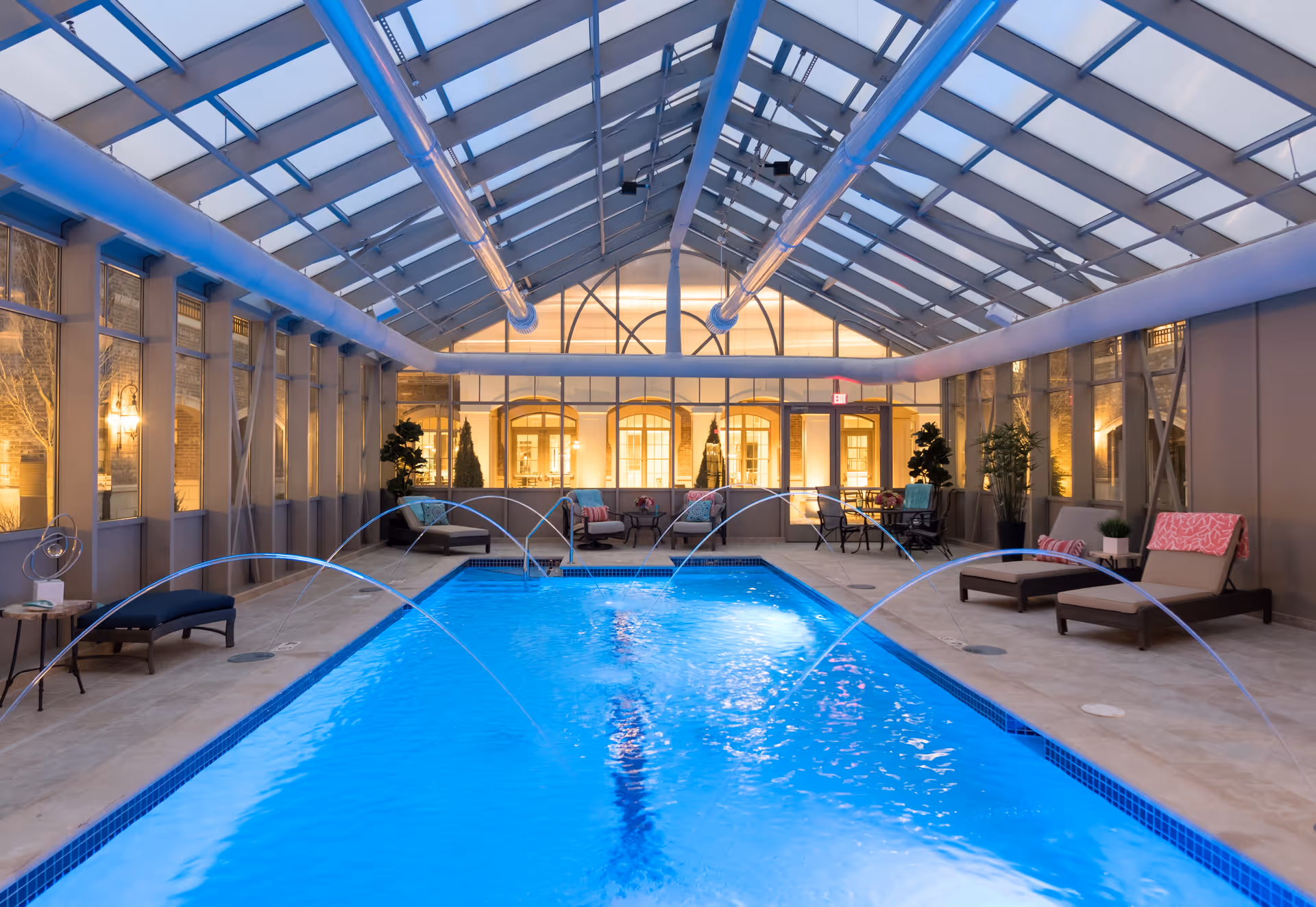 Indoor swimming pool with water jets arching over the pool, surrounded by lounge chairs and seating areas under a glass ceiling with metal beams, illuminated with warm lighting.