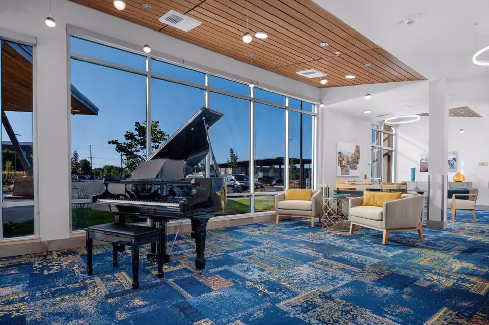 Spacious senior living lounge with a grand piano by large windows and a seating area with chairs and tables.