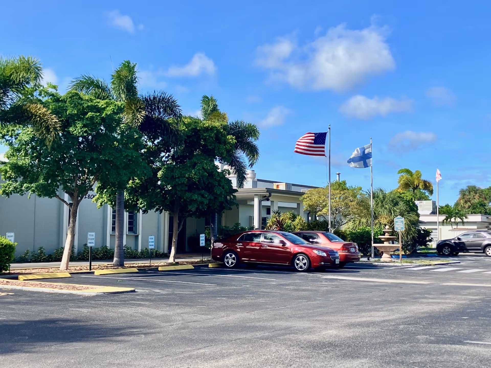 Parking lot in front of a single-story building with palm trees and other greenery. Three cars are parked, and three flags are flying on flagpoles, including the American flag and the Finnish flag. The sky is blue with some clouds.