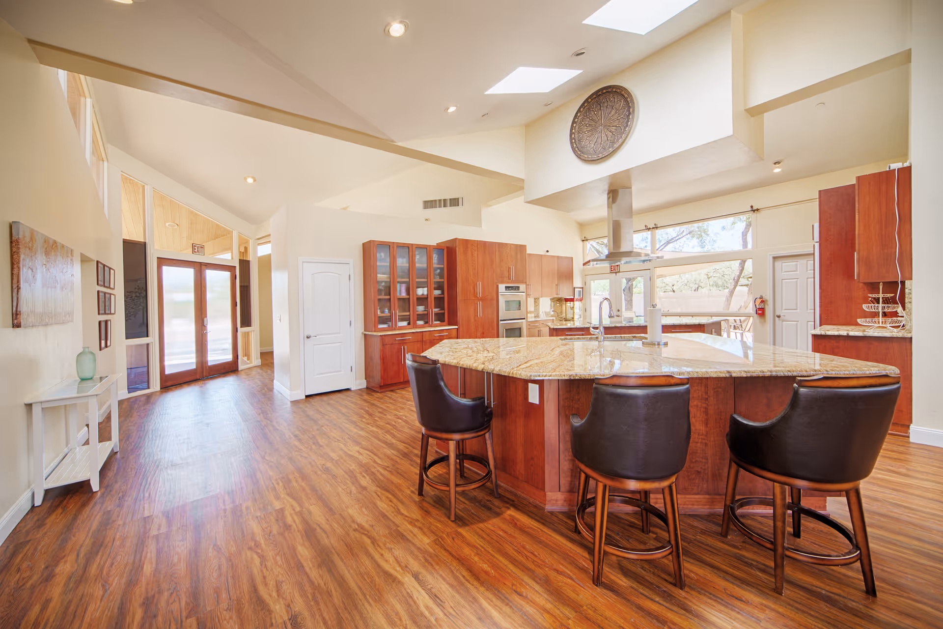 Spacious kitchen area with a large granite island countertop surrounded by three dark leather bar stools. The kitchen features wooden cabinets, built-in ovens, a modern range hood, and large windows allowing natural light to fill the room. The floor is wood, and there is a hallway with double glass doors leading outside.