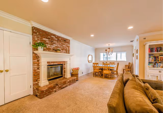 Cozy living room with brick fireplace, sofa, bookshelf, and dining area in background.