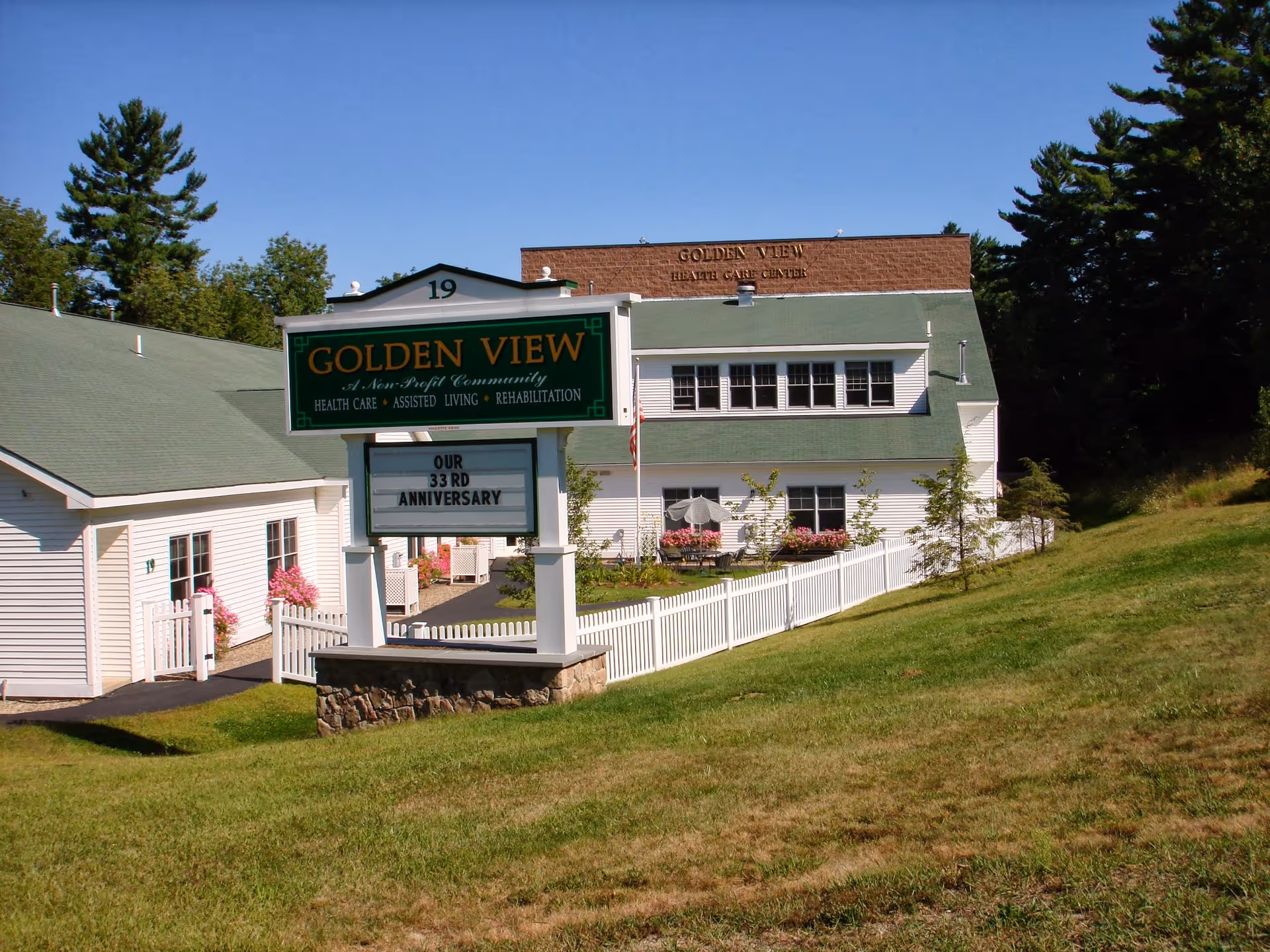 Front exterior of Golden View Health Care Center with a green-roofed building, white fence, and a large sign announcing the facility and its 33rd anniversary.