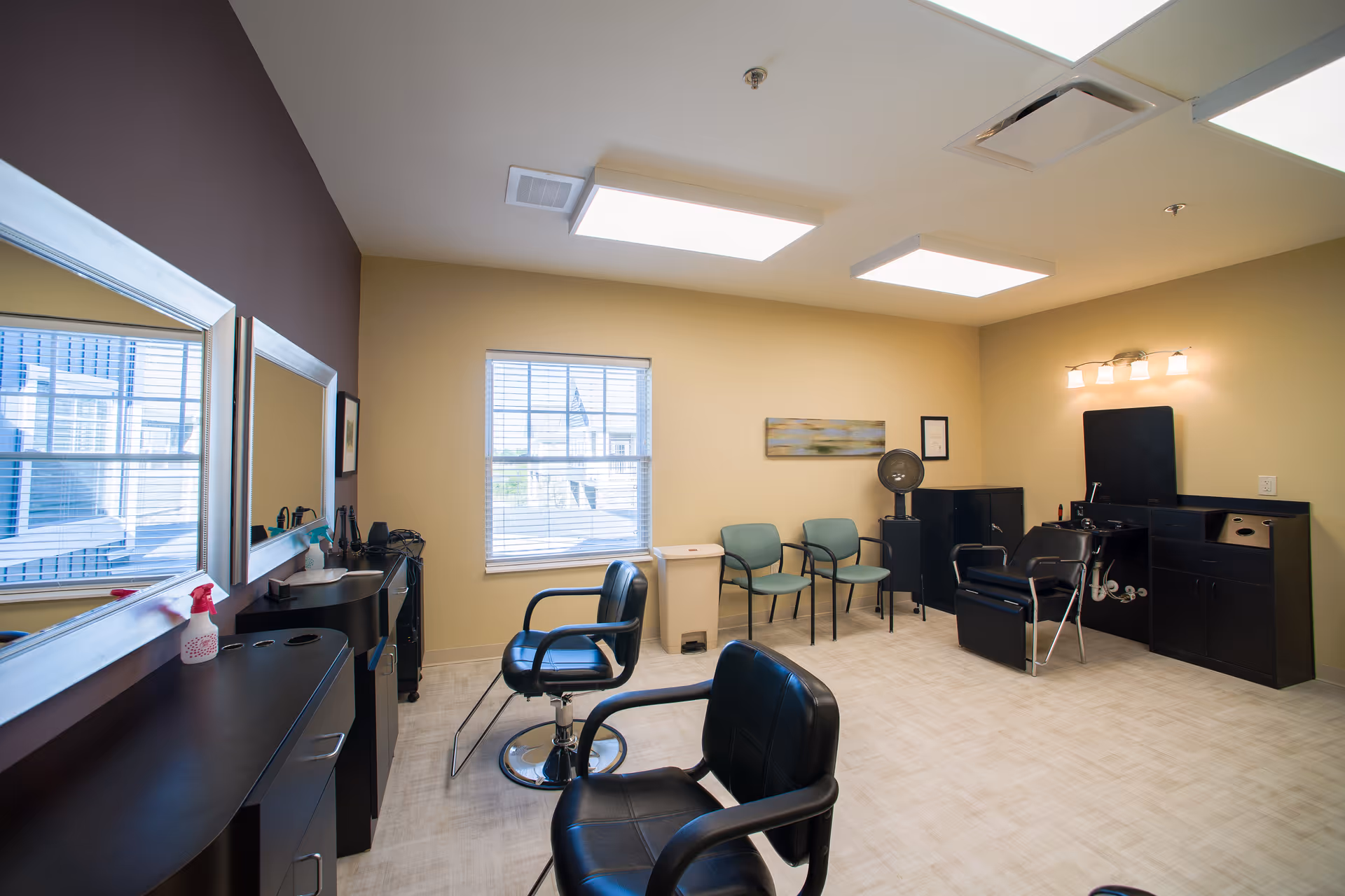 Interior view of a hair salon area in a senior living facility with black salon chairs, large mirrors on the wall, a window with blinds, two green waiting chairs, a hair dryer, and overhead fluorescent lighting.