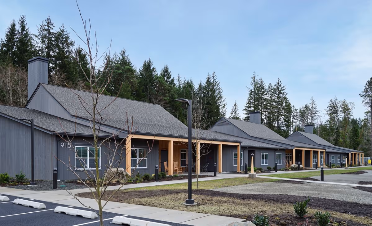 Exterior view of a single-story senior living facility building with gray siding and a sloped roof, surrounded by trees and a parking area in front. The building has multiple windows and covered porch areas with wooden posts.