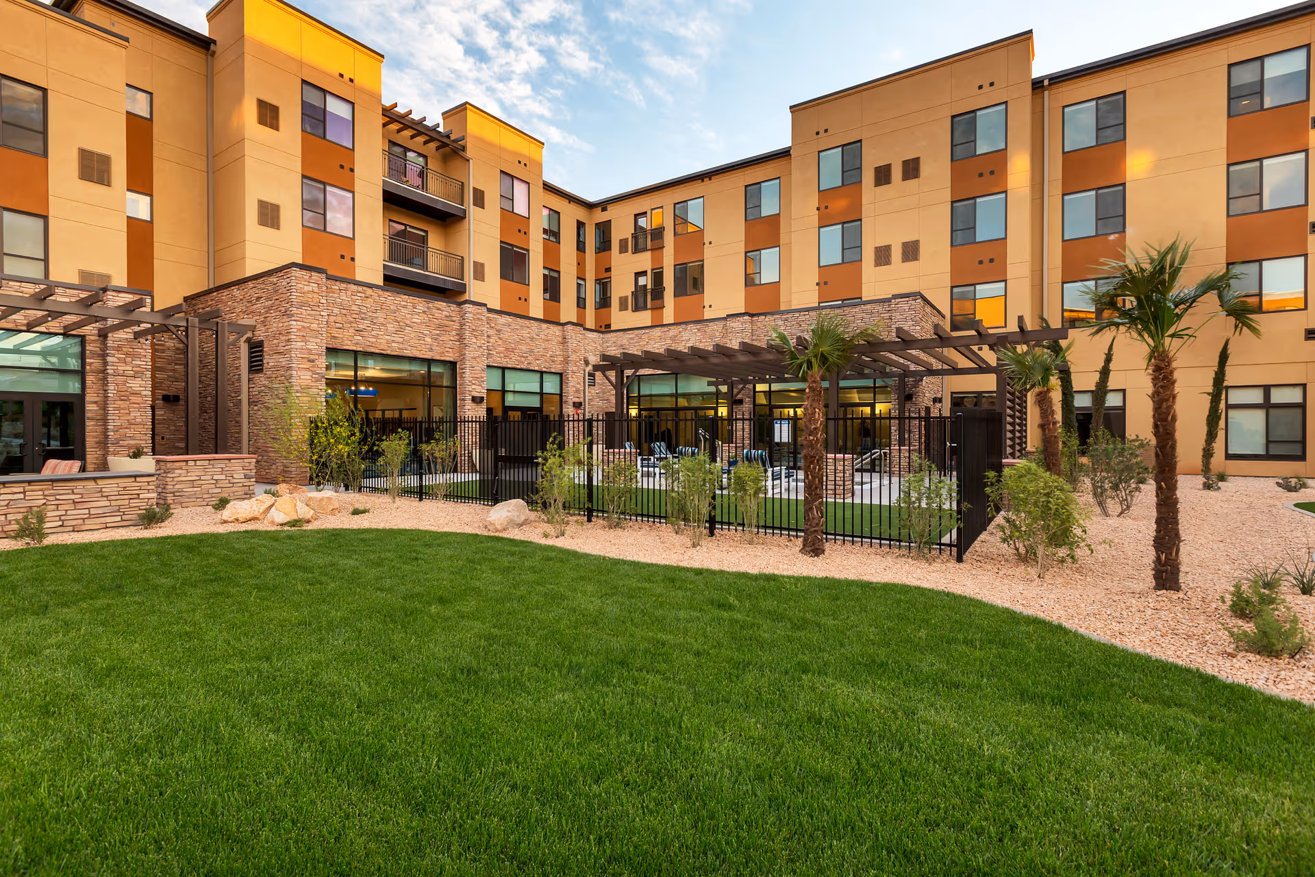 Outdoor courtyard area of a senior living facility with green grass, palm trees, and a fenced patio with seating. The building surrounding the courtyard is multi-story with a combination of stone and beige and orange exterior walls and many windows.