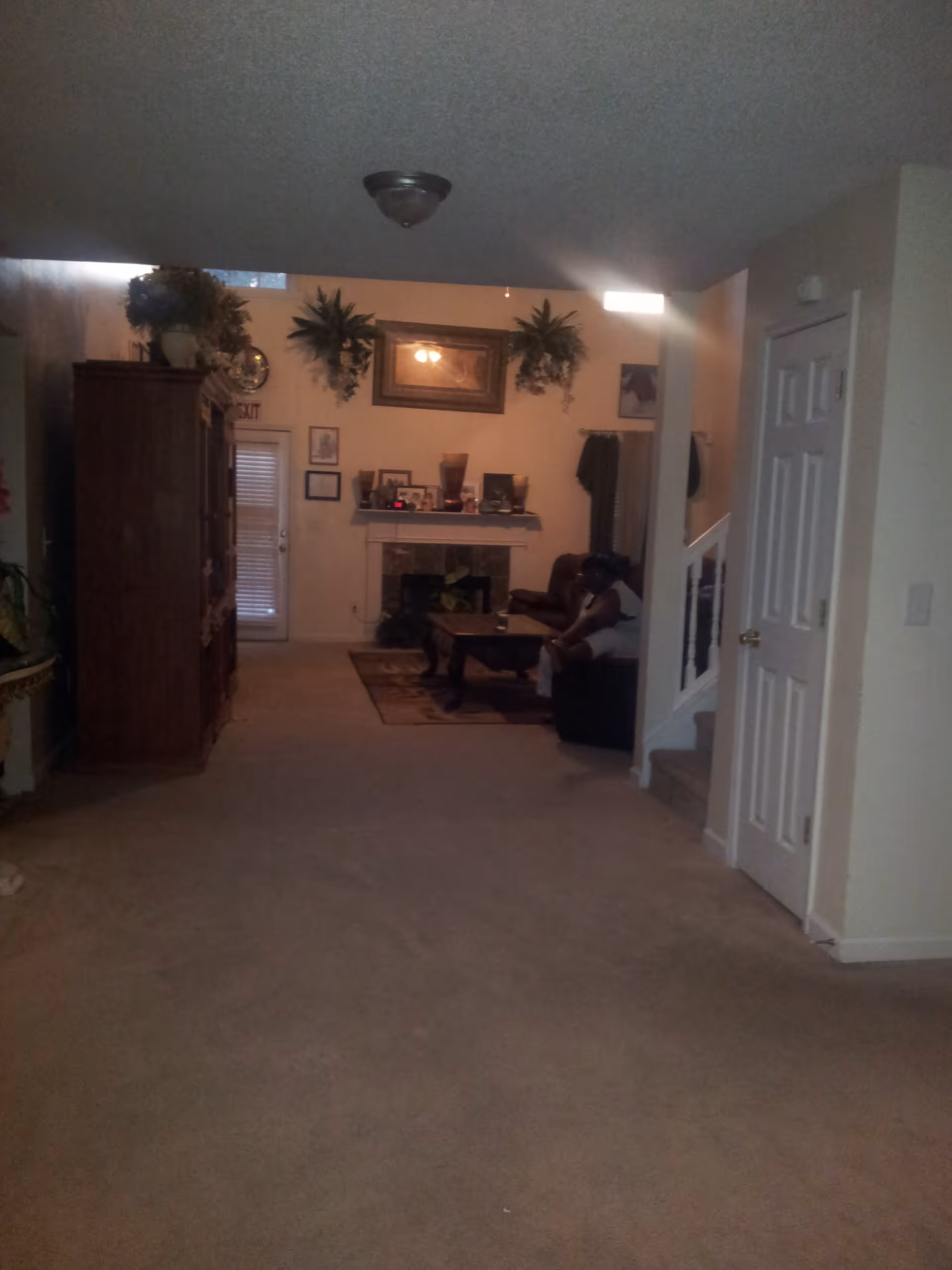 Interior view of a living room with beige carpet and walls. There is a wooden cabinet on the left, a fireplace with decorations and framed pictures above it in the center, and a person sitting on a dark sofa near a coffee table on the right. The room has some plants and a staircase with a white railing on the right side.