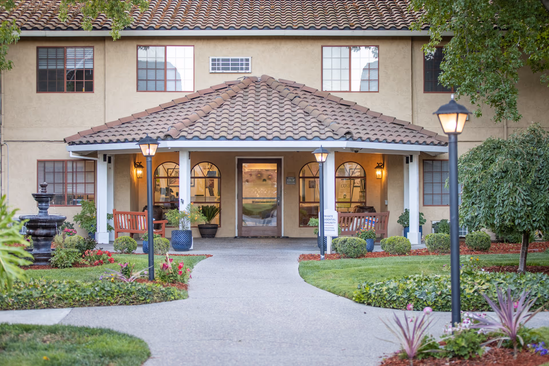 Front exterior view of a senior living facility named Cogir of Manteca with a tiled roof, a covered entrance supported by white pillars, two benches, potted plants, a pathway leading to the entrance, garden landscaping with bushes, flowers, and two lit lamp posts.