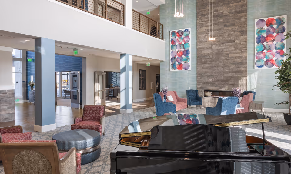 A spacious and well-lit living room area with a black grand piano in the foreground. The room features multiple seating arrangements with pink and blue armchairs, a round ottoman, and side tables with flower vases. The walls have modern art pieces with colorful circular patterns and a tall stone fireplace in the center. Large windows allow natural light to fill the space, and there is a second-floor balcony overlooking the area.