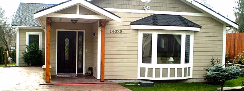 Front exterior view of a single-story house with beige siding, a dark front door with glass panels, a small covered porch supported by wooden posts, a bay window with white trim, and a well-maintained lawn with shrubs and a small tree.