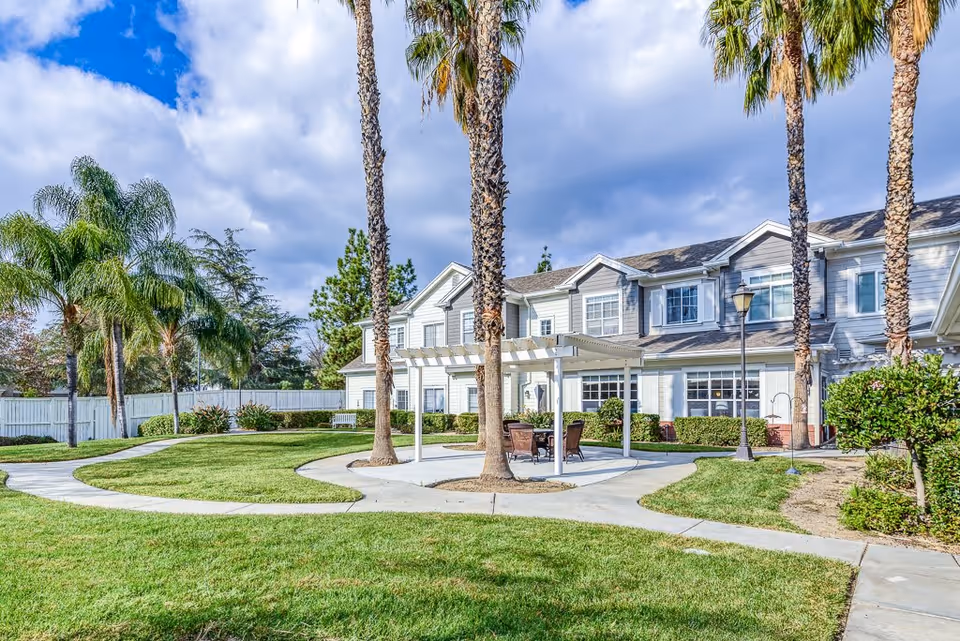 Outdoor garden area at Gardens At Northridge featuring a paved walkway, green lawn, palm trees, a white pergola with outdoor seating, and a two-story building in the background under a partly cloudy sky.