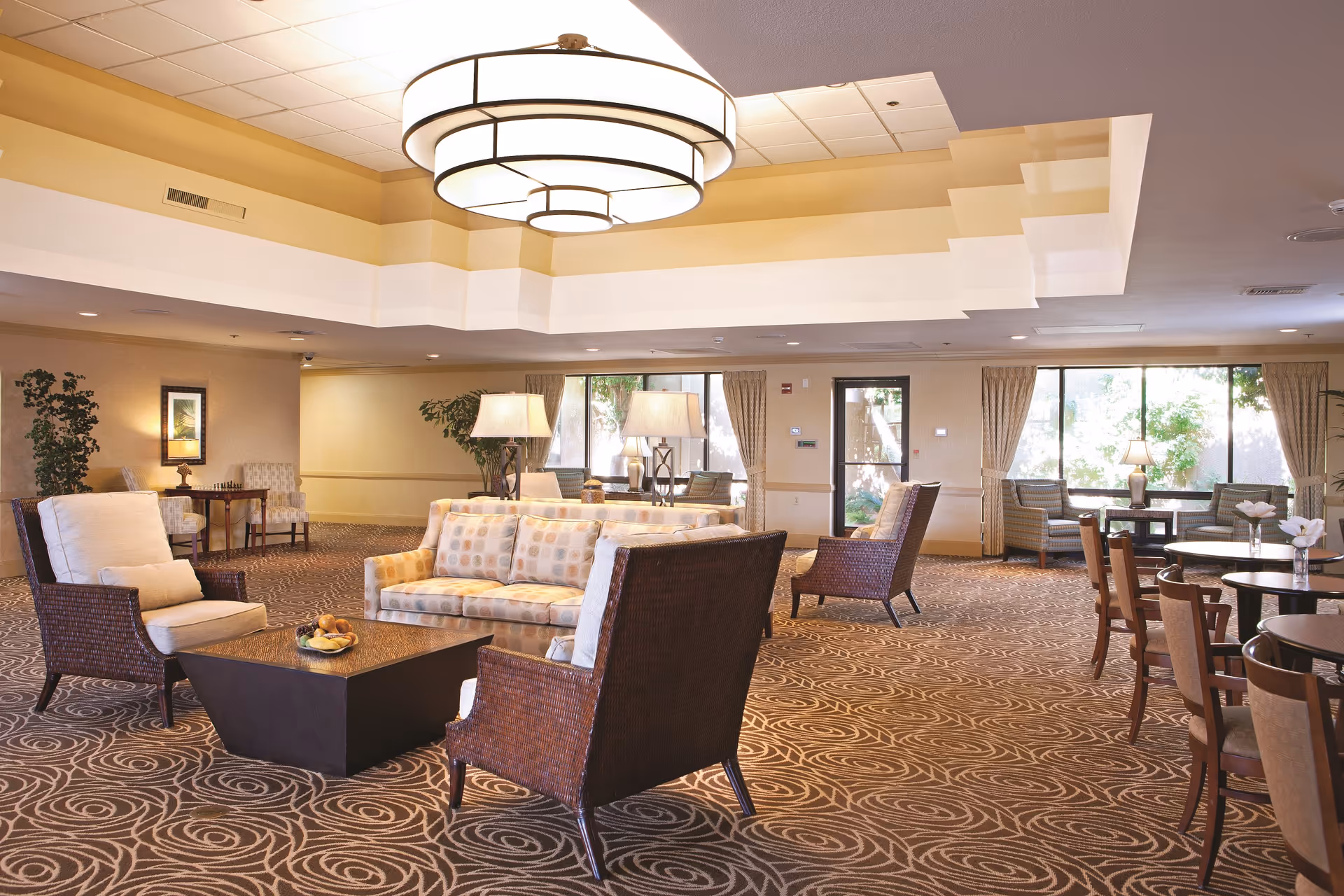 A spacious and well-lit common area in a senior living facility featuring comfortable seating including cushioned armchairs and a patterned sofa around a central coffee table. The room has large windows with curtains allowing natural light, decorative lamps, patterned carpet, and a modern ceiling light fixture. Additional seating and tables are visible in the background.