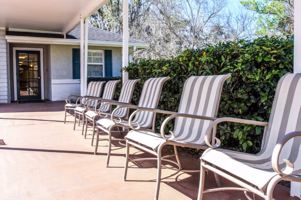 Row of striped outdoor patio chairs lined up under a covered walkway beside a building entrance and hedges.