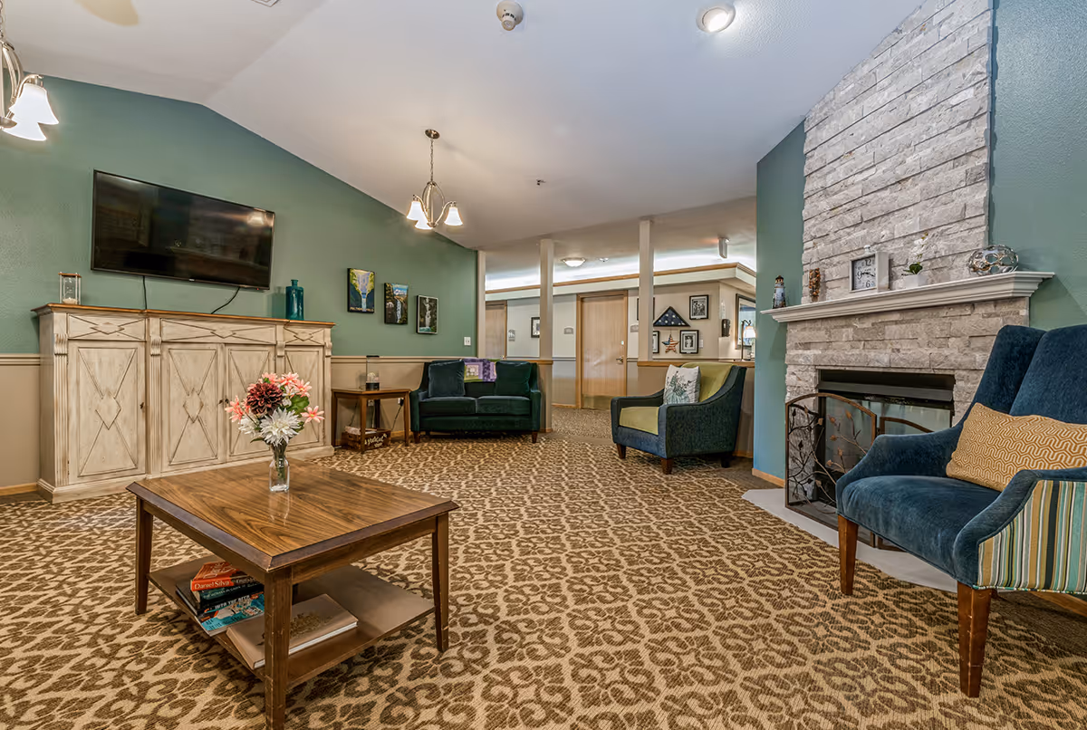 A cozy living room area with patterned carpet, a stone fireplace on the right, a blue armchair with a yellow pillow, a green armchair, and a dark green loveseat. A wooden coffee table with a vase of flowers and books underneath is in the center. On the left wall, there is a mounted flat-screen TV above a white distressed cabinet, with three small paintings hanging on the green wall. The room is softly lit with ceiling lights and has a warm, inviting atmosphere.
