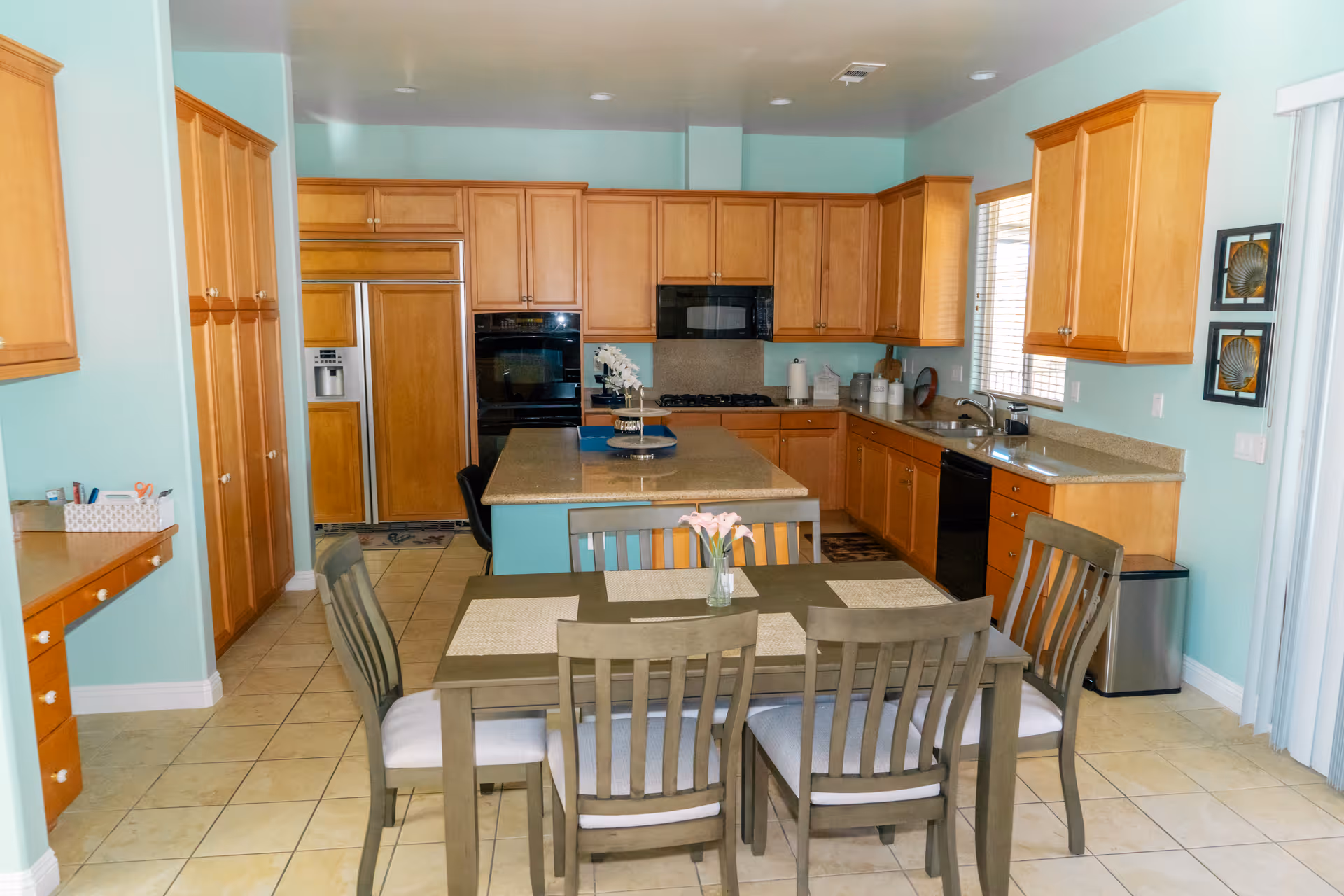A spacious kitchen with light wood cabinets, a central island, and a dining table with six chairs. The kitchen features a refrigerator, oven, microwave, dishwasher, and a sink under a window. The walls are painted light blue, and the floor is tiled. There are two framed pictures on the wall near the window.