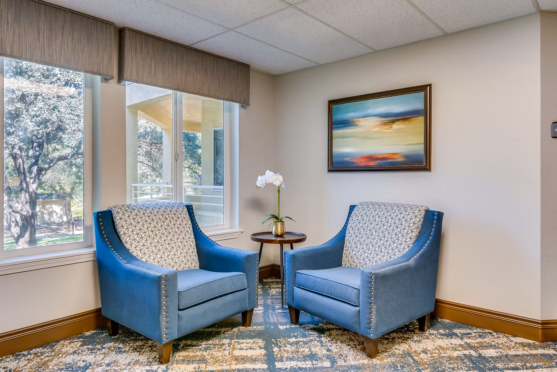 A cozy seating area with two blue upholstered armchairs featuring patterned back cushions, positioned around a small round wooden table with a white orchid plant in a gold pot. The area is next to large windows with a view of trees outside, and a colorful abstract painting hangs on the beige wall.