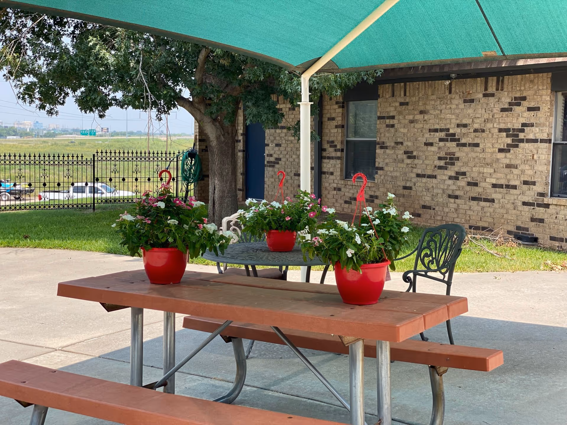 Outdoor seating area with a wooden picnic table and metal chairs under a green canopy. Three red pots with flowering plants are placed on the tables. A brick building and a tree are visible in the background, along with a black metal fence and a grassy field beyond.