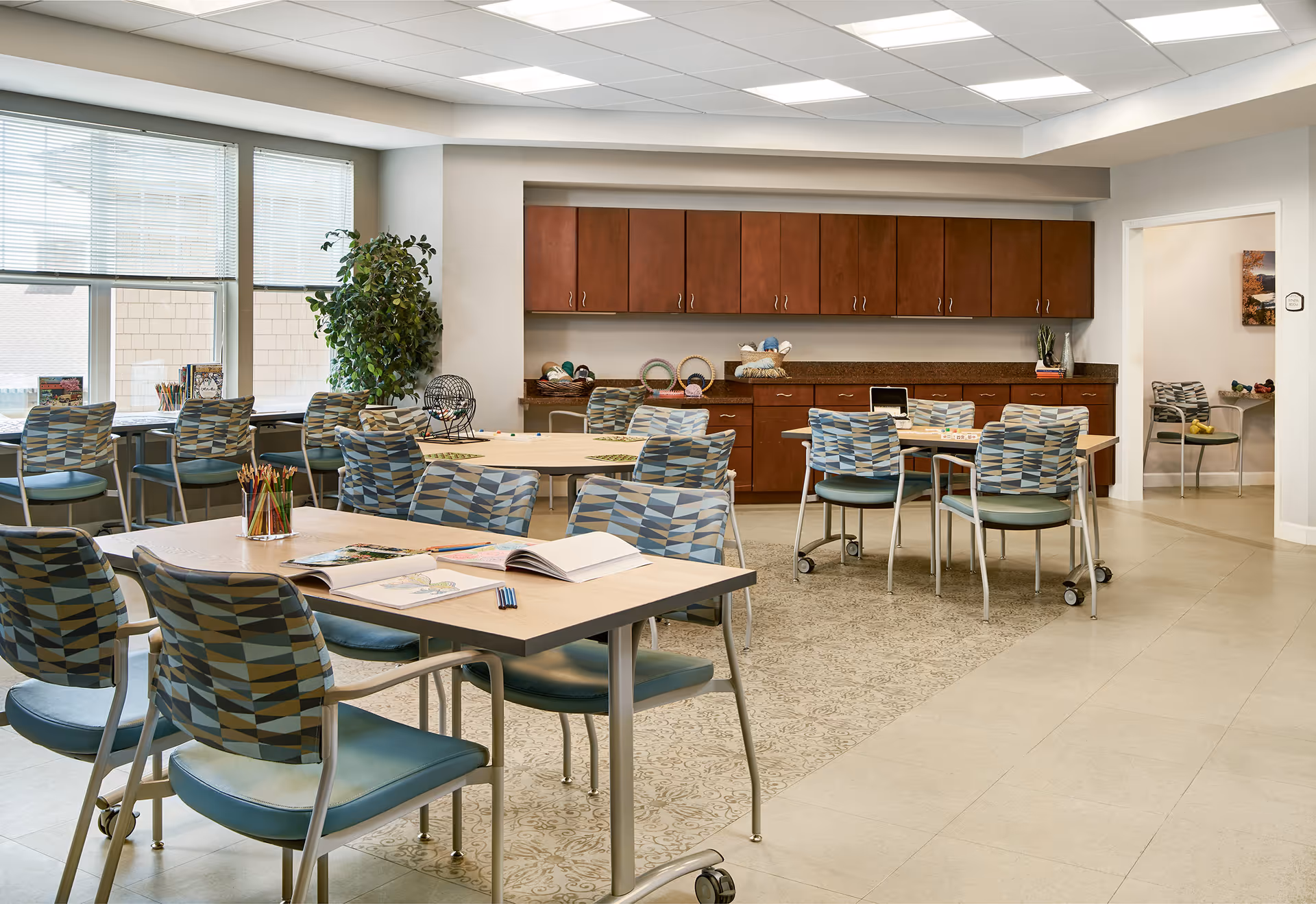 A bright and spacious activity room in a senior living facility with multiple tables and chairs arranged for group activities. The tables have coloring books, pencils, and other craft supplies. There are large windows letting in natural light, a potted plant, and wooden cabinets along the back wall with various craft materials on the countertop.