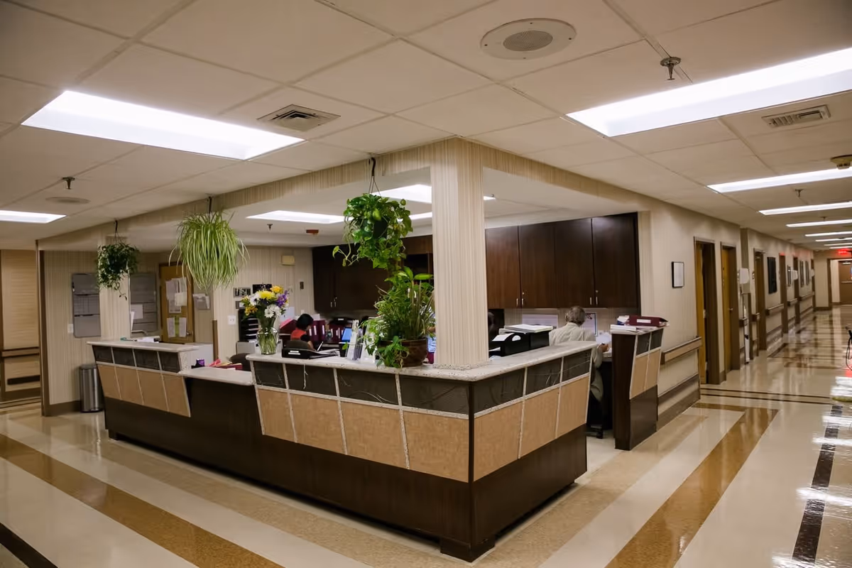 Interior view of a nursing station in a healthcare facility with a long counter, hanging plants, flowers in a vase, and staff working at computers. The area is well-lit with fluorescent ceiling lights and has a polished floor with brown and beige stripes. A hallway with multiple doors extends to the right.