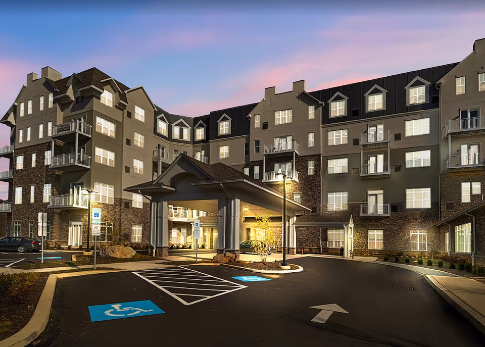 Exterior view of a multi-story senior living facility building at dusk with lit windows and a covered entrance. The parking area in front includes designated handicapped parking spaces and a driveway with an arrow pointing forward.