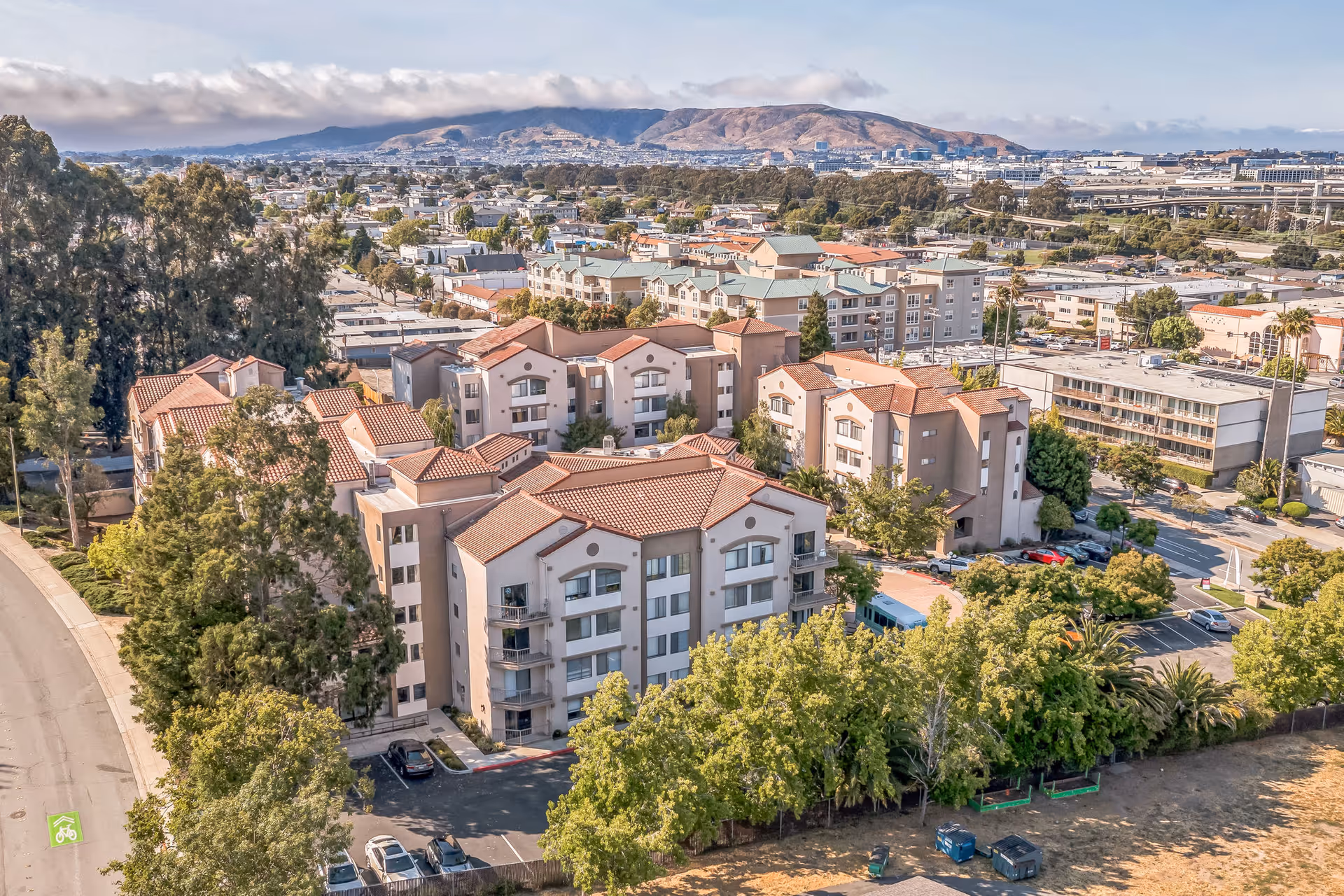 Aerial view of the Cadence Millbrae senior living complex showing multiple Mediterranean-style apartment buildings, parking areas, trees, and distant hills.