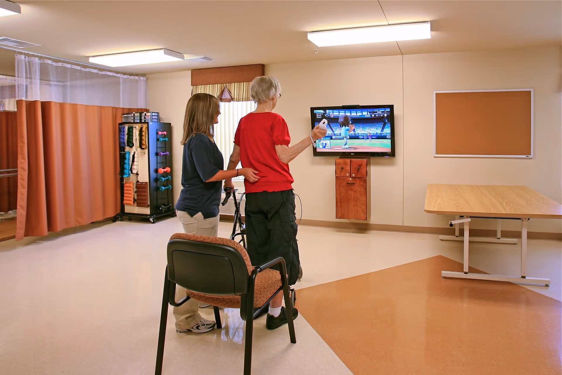 An elderly person using a walker is playing a video game on a wall-mounted TV with the assistance of a caregiver in a spacious room. The room has a chair, a table, exercise equipment, and a curtain divider.