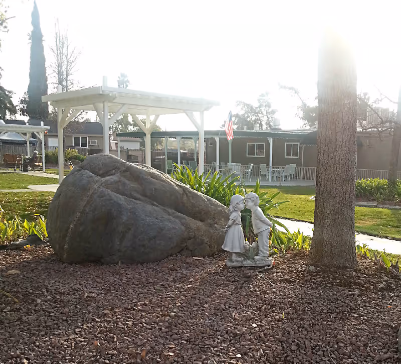 Outdoor garden with a large decorative rock, a statue of two children, pergolas, and a building with an American flag in the background.